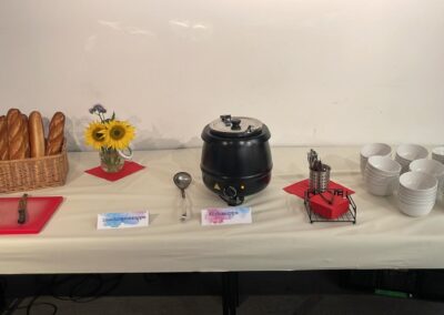 Ein gedeckter Tisch mit einer Suppe, frischen Brötchen und buntem Blumenbouquet. Der Tisch ist dekoriert mit Tellern und Besteck.